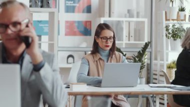 Güzel esmer iş kadını şık günlük kıyafetleri ve gözlükleriyle açık uzay ofisinde oturuyor, laptopta mesaj atıyor ve iş günü boyunca düşünüyor.