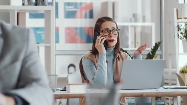 穿着智能休闲装 戴着眼镜 坐在办公桌前 一边用手机讲话 一边在露天办公室工作的女商人的追踪照片 — 图库视频影像