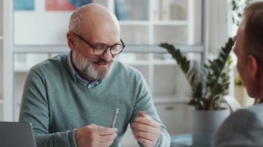 Neşeli kıdemli iş adamının, erkek meslektaşıyla konuşurken ve ofiste otururken notlar alırken çekilmiş bir fotoğrafı.