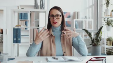 Çekici esmer iş kadını şık günlük kıyafetleri ve gözlükleriyle ofiste oturuyor ve web konferansı sırasında ya da video blogu kaydederken kamerada bir şeyler anlatıyor.