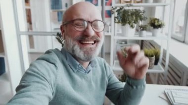 Ofisteki iş yerinde kablosuz kulaklıklı kıdemli işadamı kameraya bakıyor, akıllı telefondan görüntülü telefon aracılığıyla sohbet ederken gülümsüyor ve konuşuyor.