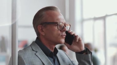 Formalite ve gözlüklü ciddi orta yaşlı işadamının yakın plan çekimleri ofiste cam duvarın yanında duruyor ve cep telefonuyla konuşuyor.