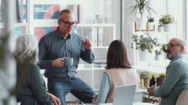 Orta yaşlı iş adamı elinde çay fincanı tutarak ofiste brifing verirken karışık yaştaki meslektaşlarıyla konuşuyor.