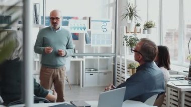 Cam ofis duvarındaki posterlere işaret eden kıdemli iş adamı sunumu açıklıyor ve takım toplantısı sırasında iş arkadaşlarıyla tartışıyor.