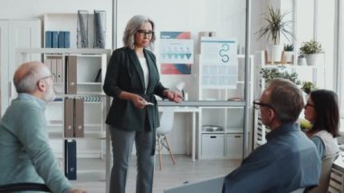 Güzel kıdemli iş kadını cam duvardaki posterlere işaret ediyor ve ofis toplantısı sırasında karışık yaştaki meslektaşlarına proje hakkında sunum yapıyor.