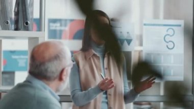 Eğitimsel seminer sırasında çalışanlara öğretmenlik yaparken cam ofis duvarına asılmış posterlerdeki bilgileri açıklayan el kamerası görüntüsü.