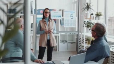 Genç esmer iş kadını cam duvardaki posterlere işaret ediyor ve ofis toplantısında sunum yaparken karışık yaşlardaki profesyonellere bilgi aktarıyor.