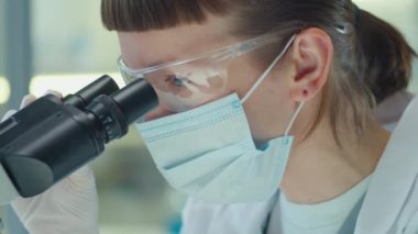 Laboratuvarda çalışırken mikroskop kullanarak koruyucu maskeli ve gözlüklü genç kadın bilim adamının yakın plan fotoğrafını çek.