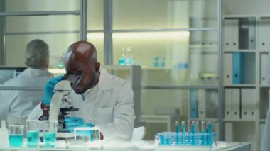 Genç Afrikalı Amerikalı bilim adamı laboratuvarda mikroskop kullanırken Kafkas kadın meslektaşı ona doğru yürüyor, dijital tablette bir şeyler gösteriyor ve konuşuyor.