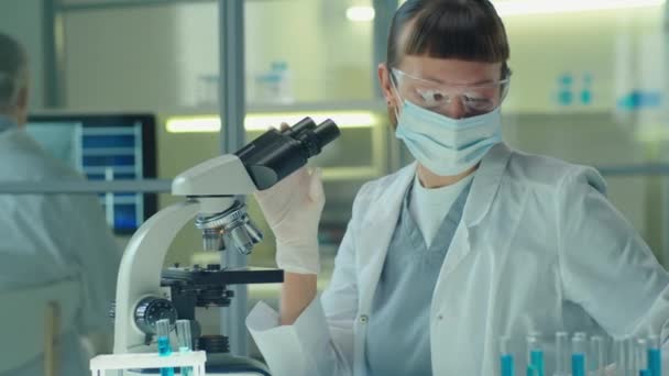 研究室でワクチン研究をしながら顕微鏡を用いた顔の保護マスク 手袋の女性科学者 — ストック動画