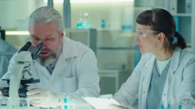 Beyaz önlüklü ve koruyucu eldivenli kıdemli beyaz bilim adamı mikroskoptan bakıyor ve laboratuvarda dijital tablet kullanırken genç bayan meslektaşıyla konuşuyor.