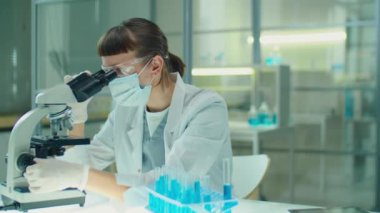 Koruyucu maskeli, gözlüklü ve eldivenli kadın kimyager laboratuarda test tüpleriyle çalışırken mikroskoptan bakıyor.
