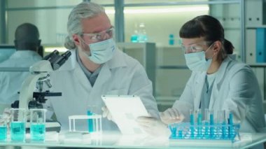 Beyaz önlüklü, koruyucu maskeli ve eldivenli yetişkin bir beyaz bilim adamı mikroskop kullanıyor ve laboratuvarda dijital tabletle çalışırken genç bayan meslektaşıyla konuşuyor.