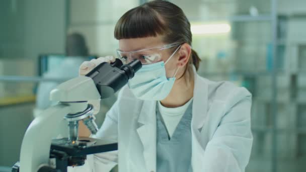 Vrouwelijke Laboratoriummedewerker Beschermend Masker Bril Handschoenen Kijkend Door Microscoop Tijdens — Stockvideo