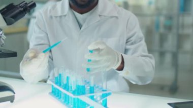 Beyaz önlüklü ve eldivenli Afrikalı Amerikalı erkek bilim adamının test tüpünü askıdan alıp laboratuarda çalışırken içine mavi kimyasalları dökerken çekimini yukarı kaldır.