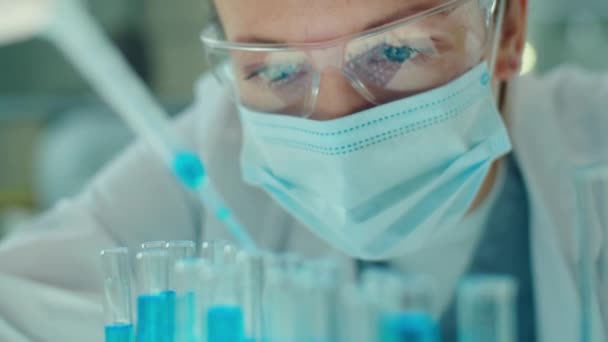 실험실에서 미생물학 실험을 보호용 마스크와 안경을 과학자가 시험관에 파란색 액체를 — 비디오
