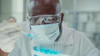 Koruyucu maskeli, gözlüklü ve eldivenli Afrikalı Amerikalı laboratuar çalışanının mikrobiyoloji araştırması yaparken deney tüplerine pipetle mavi kimyasallar dökerken seçici odak noktası.