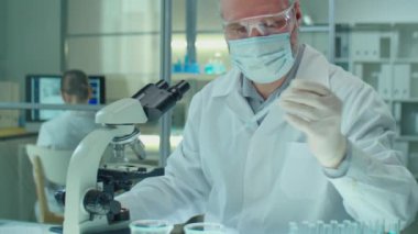 Koruyucu maskeli, eldivenli ve gözlüklü kıdemli Kafkas kimyageri cam parçasına numune yerleştiriyor ve laboratuvarda çalışırken mikroskop altında inceliyor.