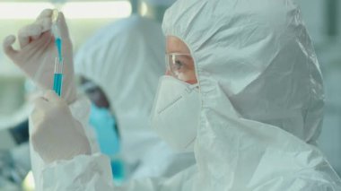 Koruyucu giysi, gözlük ve maske takan kadın laboratuvar çalışanı Omikron 'a karşı aşı üzerinde çalışırken pipetle test tüpüne mavi sıvı dolduruyor.