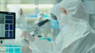 Laboratuvarda kovid aşısı üzerinde çalışırken koruyucu giysili ve maskeli kimyager deney tüplerine pipetle mavi sıvıyı döküyor.