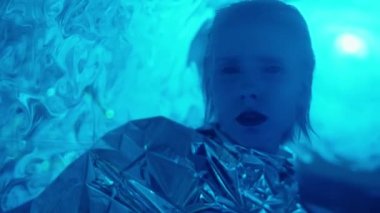 Stüdyo resmi, siyah gözlü ve rujlu, folyo elbise giyen, kameraya mavi lazer ışığında poz veren uzaylı kadın.