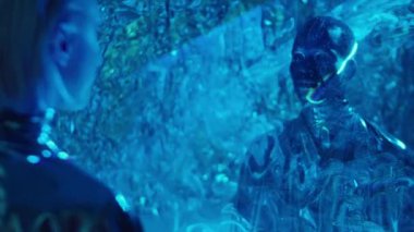 Yaratıcı makyajlı, gümüş cüppeli uzaylı bir adamın omzunun üstünden. Stüdyoda mavi ışıklı projeksiyonla duruyor ve kadına bakıyor.