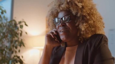 Neşeli Afrikalı Afrikalı Amerikalı iş kadını gece geç saatlerde ofiste çalışırken cep telefonuyla sohbet ediyor.