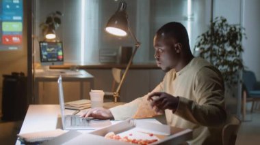 Afro-Amerikalı iş adamı laptopta çalışıyor ve bütün gece ofis masasında pizza yiyor.