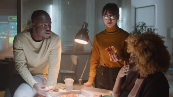 多民族の男性と女性の同僚のチームは 深夜にオフィスで一緒に働いている間 ピザを食べて ビジネスについて話しています — ストック動画