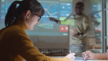 Afrika kökenli Amerikalı bir adamın sunumunu dinleyen ve yatırım şirketinin ofisinde gece iş görüşmeleri yaparken not alan çok ırklı kadın meslektaşların net bir fotoğrafı.