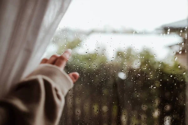 Window view with raindrops with blurred hand in sweatshirt. Atmospheric cozy shots of rainy mood.
