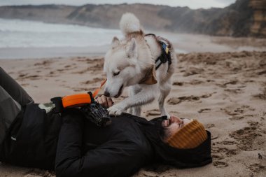 Beyaz Samur Sibirya Husky bir adam kumların üzerinde yatarken sahibiyle oynuyor. Avustralya 'da Büyük Okyanus Yolu' nda bir sahilde bulutlu bir gün. Köpeğiyle oynayan bir adamla..