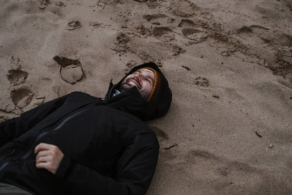 Smiling man in a black raincoat lying on a sand in winter. Cloudy day on a beach along Great Ocean Road in Australia with a man lying on a sand.