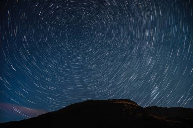 Ayışığında dağların üstündeki yıldızlı gökyüzünde dairesel izler şeklinde dönen yıldızların izleriyle soyut bir gece manzarası.
