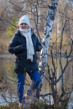 Sonbahar günü. Şapkalı, örgülü ve kalın kazaklı bir kadın. Gölün kıyısında duruyor ve bu yıl güneşin son ışınlarının tadını çıkarıyor. Arka planda Birch korusu.