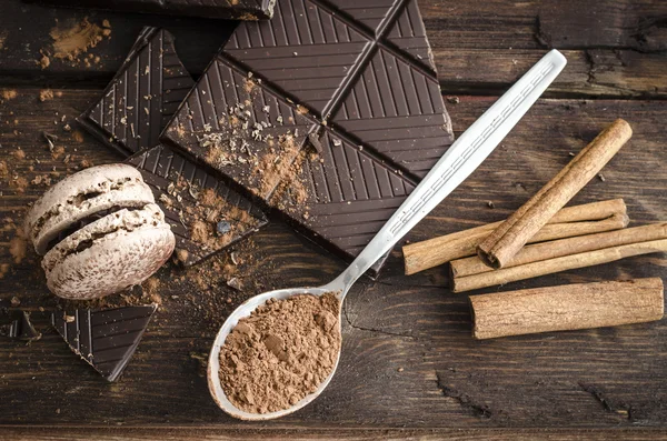 Mörk choklad, kakaopulver, franska macaron och kanelstänger på träbord Stockfoto