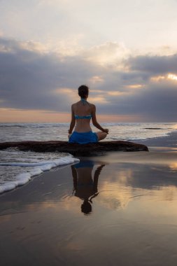 Gün batımı yogası. Lotus pozuyla taşın üzerinde oturan beyaz kadın. Padmasana. Eller Gyan Mudra 'da. Bali 'de bir sahil. Arkadan bak. Uzayı kopyala Yoga inzivası. Su yansıması. Kişisel bakım konsepti. Zen hayatı.