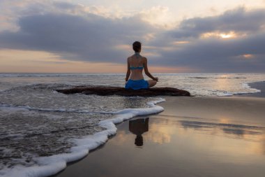 Gün batımı yogası. Lotus pozuyla taşın üzerinde oturan beyaz kadın. Padmasana. Eller Gyan Mudra 'da. Bali 'de bir sahil. Arkadan bak. Uzayı kopyala Yoga inzivası. Su yansıması. Sağlıklı bir yaşam tarzı. Zen hayatı.