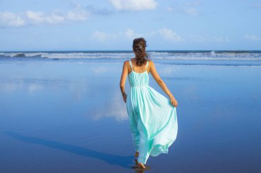 Genç bir kadın boş sahilde yalınayak yürüyor. Tam vücut portresi. İnce beyaz kadın, uzun elbise giyiyor. Arkadan bak. Yaz güneşi. Mavi gökyüzü. Asya 'da tatil. Seyahat konsepti. Bali, Endonezya