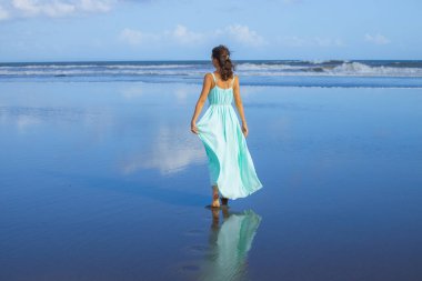 Genç bir kadın boş sahilde yalınayak yürüyor. Tam vücut portresi. İnce beyaz kadın, uzun elbise giyiyor. Arkadan bak. Yaz güneşi. Mavi gökyüzü. Asya 'da tatil. Seyahat konsepti. Bali, Endonezya