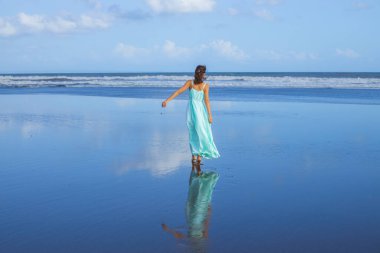 Genç bir kadın boş sahilde yalınayak yürüyor. Tam vücut portresi. İnce beyaz kadın, uzun elbise giyiyor. Arkadan bak. Su yansıması. Mavi gökyüzü. Asya 'da tatil. Seyahat konsepti. Bali, Endonezya