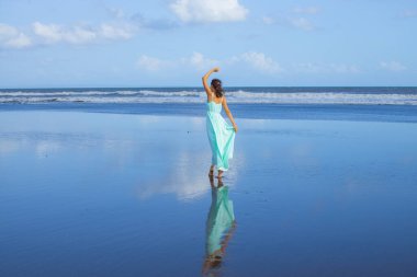 Genç bir kadın boş sahilde yalınayak dans ediyor. Tam vücut portresi. İnce beyaz kadın, uzun elbise giyiyor. Arkadan bak. Su yansıması. Mavi gökyüzü. Asya 'da tatil. Seyahat konsepti. Bali, Endonezya