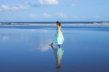 Genç bir kadın boş kumsalda yalınayak dans ediyor. Tam vücut portresi. İnce beyaz kadın, uzun elbise giyiyor. Su yansıması. Mavi gökyüzü. Asya 'da yaz tatili. Seyahat konsepti. Bali, Endonezya