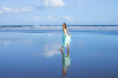 Boş sahilde çıplak ayakla yürüyen mutlu bir kadın. Tam vücut portresi. İnce beyaz kadın, uzun elbise giyiyor. Su yansıması. Mavi gökyüzü. Asya 'da yaz tatili. Seyahat konsepti. Bali, Endonezya