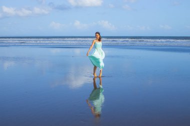 Genç bir kadın boş kumsalda yalınayak dans ediyor. Tam vücut portresi. İnce beyaz kadın, uzun elbise giyiyor. Su yansıması. Mavi gökyüzü. Asya 'da yaz tatili. Seyahat konsepti. Bali, Endonezya
