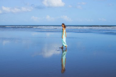 Boş sahilde çıplak ayakla yürüyen mutlu bir kadın. Tam vücut portresi. İnce beyaz kadın, uzun elbise giyiyor. Su yansıması. Mavi gökyüzü. Asya 'da yaz tatili. Seyahat konsepti. Bali, Endonezya