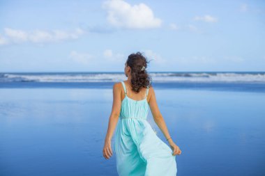 Genç bir kadın boş kumsalda yürüyor. İnce beyaz kadın, uzun elbise giyiyor. Arkadan bak. Yaz güneşi. Mavi gökyüzü. Asya 'da tatil. Seyahat konsepti. Bali, Endonezya