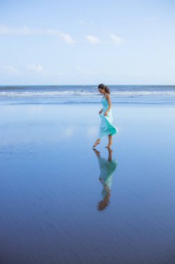Genç bir kadın boş kumsalda yalınayak yürüyor. Tam vücut portresi. İnce beyaz kadın, uzun elbise giyiyor. Su yansıması. Mavi gökyüzü. Asya 'da yaz tatili. Seyahat konsepti. Bali, Endonezya