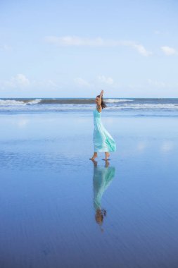 Genç bir kadın boş sahilde yalınayak yürüyor. Tam vücut portresi. İnce beyaz kadın, uzun elbise giyiyor. Ufuk çizgisi. Su yansıması. Eller yukarı! Asya 'da tatil. Seyahat konsepti. Bali