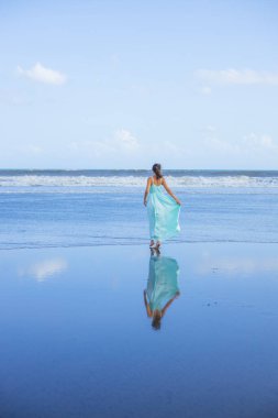 Genç bir kadın boş sahilde yalınayak yürüyor. Tam vücut portresi. İnce beyaz kadın, uzun elbise giyiyor. Arkadan bak. Su yansıması. Mavi gökyüzü. Asya 'da tatil. Seyahat konsepti. Bali, Endonezya